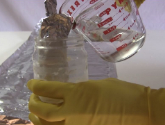 The homemade electrolytic capacitor in a container with more electrolyte being poured in.